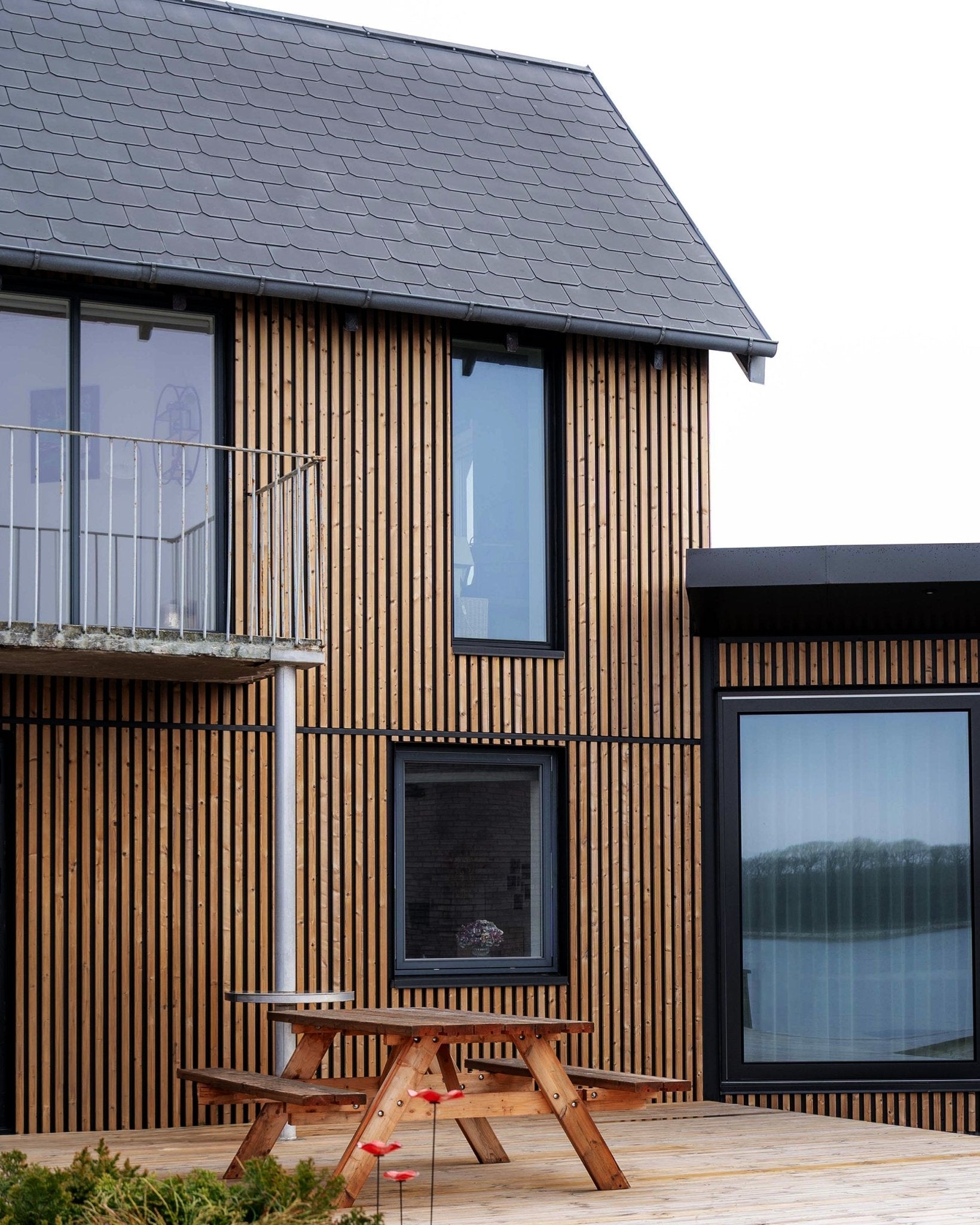 Maison moderne avec panneaux de façade WoodUpp Aluwood en Brown huilé, avec fenêtres large, un balcon et une terrasse avec table de pique-nique en bois.